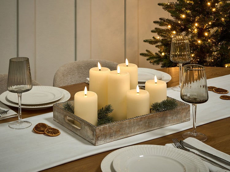 Festlich gedeckter Tisch mit LED-Kerzen im Holztablett, Geschirr und Gläsern vor einem Weihnachtsbaum.