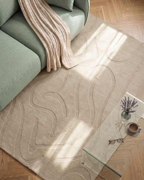 Wohnzimmer mit beigem Teppich mit abstraktem Wellenmuster, grünem Sofa, gestrickter Decke, Glastisch und Parkettboden aus der Vogelperspektive.