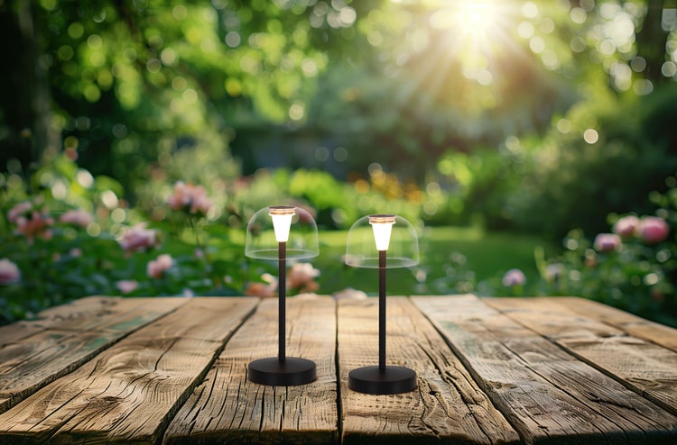 Zwei Tischleuchten mit transparentem Schirm auf einem Holztisch im Garten.
