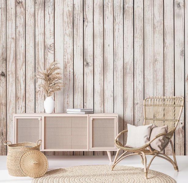 Vliestapete in heller, verwitterter Holzoptik mit vertikalen Planken in einem Raum mit Rattan-Sideboard, Sessel und Jute-Teppich.