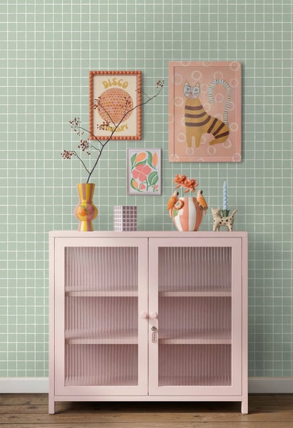 Rosa Metallschrank mit Riffelglastüren vor einer grünen Gittertapete, dekoriert mit Vasen und gerahmten Illustrationen an der Wand.