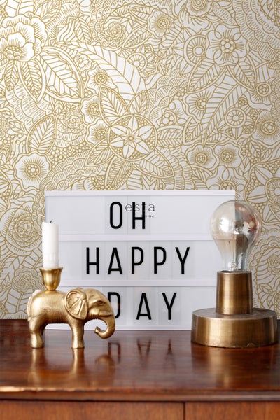 Dekoratives Stillleben mit gemusterten Tapeten, einer Leuchtbox mit der Aufschrift 'Oh Happy Day', einer Kerze in einem Elefantenständer und einer Glühbirne auf einem Tisch