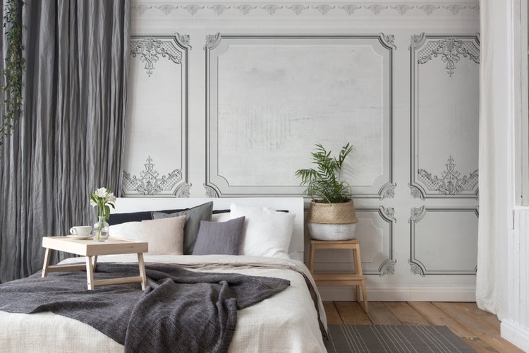 Helles Schlafzimmer mit Fototapete in dekorativer Wandpanel-Optik, einem gemachten Bett, Holzmöbeln und Zimmerpflanzen.