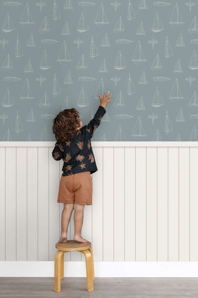 Kind steht auf einem Hocker vor einer Tapete mit Segelboot Muster und weißer Lambrisverkleidung im Kinderzimmer