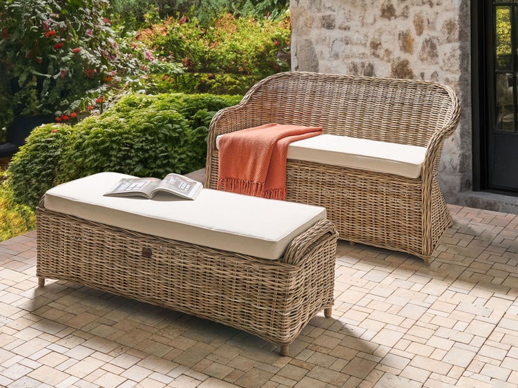 Geflochtenes Gartenmöbel-Set mit Sofa und Bank sowie hellen Polstern auf einer Terrasse mit Natursteinmauer und Pflanzen.