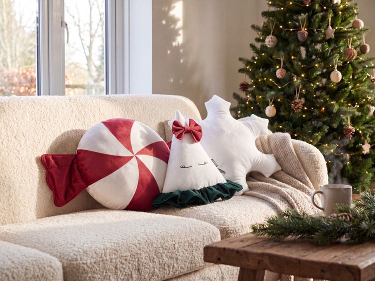 Weihnachtliche Dekokissen in Form von Bonbon, Tannenbaum und Schneeflocke auf einem hellen Sofa vor einem geschmückten Tannenbaum.