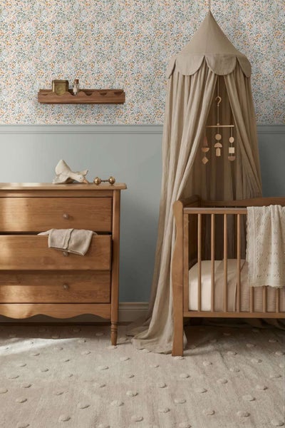 Ein Kinderzimmer mit Blumenmustertapete, Holzkommode, Gitterbett mit Betthimmel und einem strukturierten Teppich in gemütlicher Atmosphäre.