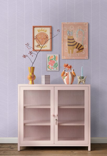 Lila Tapete mit weißem Fischgrätenmuster hinter einer rosa Vitrine mit geriffeltem Glas und bunten Dekorationsartikeln.