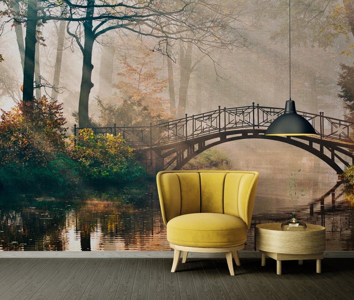 Fototapete mit nebligem Waldmotiv und Brücke, gelber Polstersessel, runder Holzbeistelltisch und schwarze Hängelampe vor dunklem Holzboden.
