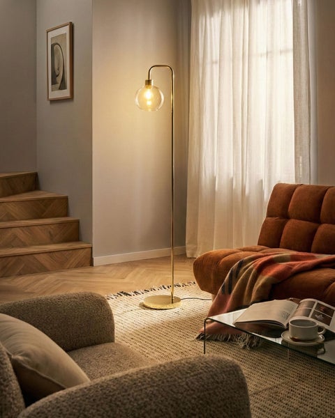 Moderne Stehlampe aus Gold mit gebogenem Arm und Glaskugelschirm in einem Wohnzimmer mit braunem Sofa und Fischgrät-Treppe.