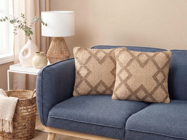 Zwei quadratische Dekokissen mit geometrischem Rautenmuster auf einem Sofa mit beigestelltem Beistelltisch, Tischlampe und geflochtenem Korb.