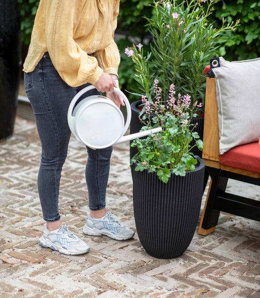 Eine Person gießt Pflanzen in einem hohen schwarzen geriffelten Pflanzgefäß mit einer weißen Gießkanne auf einer gepflasterten Terrasse.