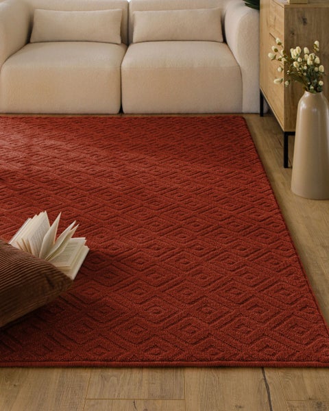 Roter Teppich mit geometrischem Rautenmuster in einem hellen Wohnzimmer auf Holzboden, neben einem Sofa und einem Kissen.