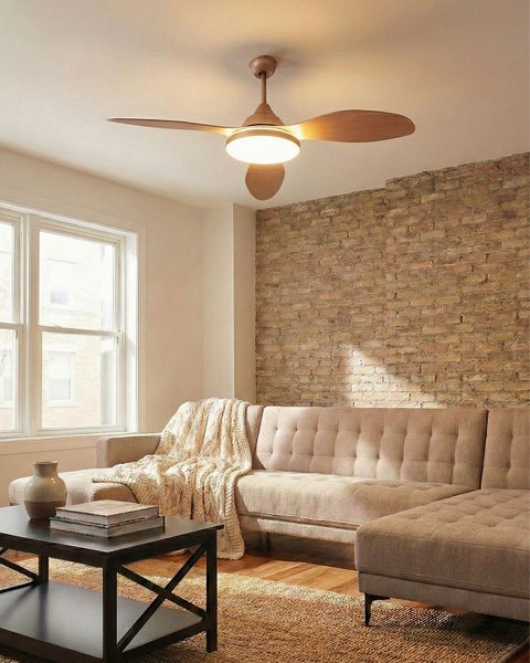 Moderner Deckenventilator mit hölzernen Flügeln und runder Leuchte in hellem Wohnzimmer mit Ziegelwand und Polster-Ecksofa.