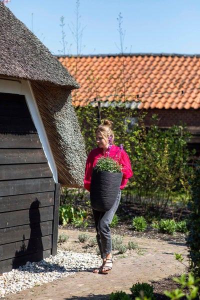 Eine Frau trägt einen großen, gerillten Pflanzkübel mit einer Pflanze auf einem Gartenweg neben einem Gebäude mit Reetdach.