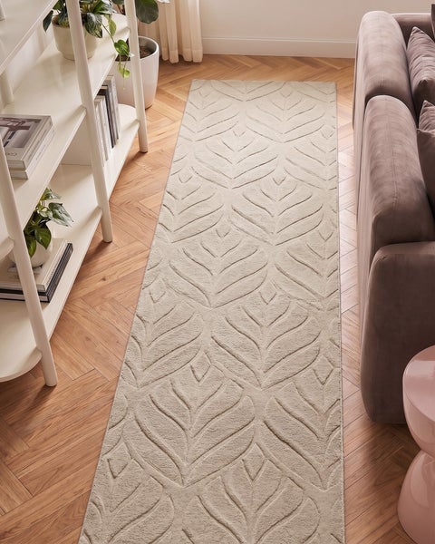 Rechteckiger Teppichläufer mit geprägtem Blattmuster auf Parkettboden in einem hellen Wohnzimmer neben einem weißen Regal und einem Sofa.