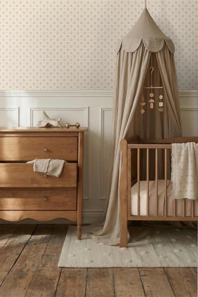 Eingerichtetes Kinderzimmer mit Holzkommode, Gitterbett aus Holz mit Baldachin, gemusterter Tapete und klassischen Wandpaneelen.