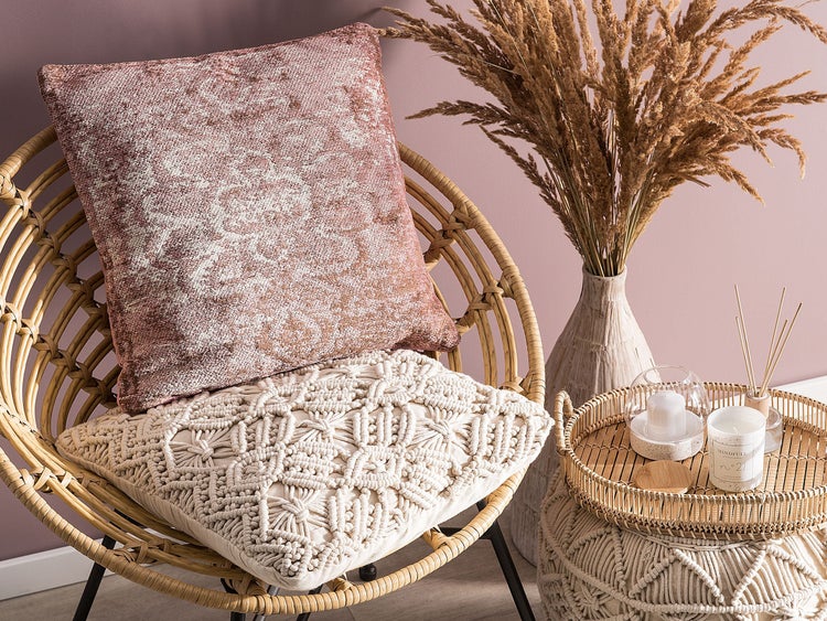 Heller Raum mit Rattansessel, einem rosa Zierkissen mit Muster und einem beigen Makramee-Kissen neben einer Vase mit Trockenblumen.