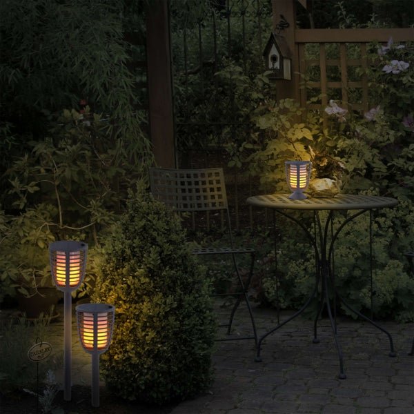 Gartenszene mit Holztisch und zwei Gartenfackeln mit Erdspieß und einer Tischgartenfackel.