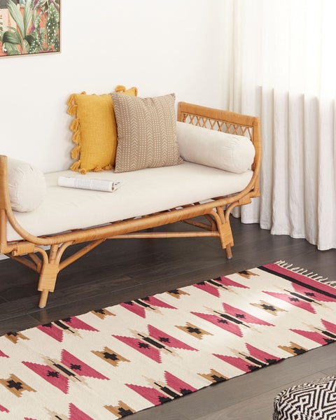 Rattanbank mit hellen Polstern und Zierkissen auf einem gemusterten Teppich in einem hellen Wohnraum mit dunklem Boden.