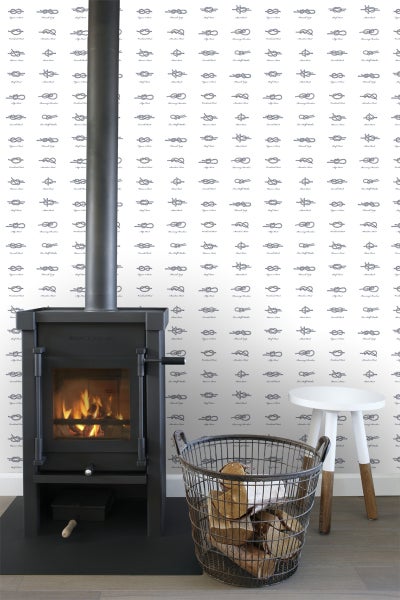 Wohnzimmer mit Kaminofen, Brennholzkorb und Hocker vor einer Wand mit Knotenmuster-Tapete.