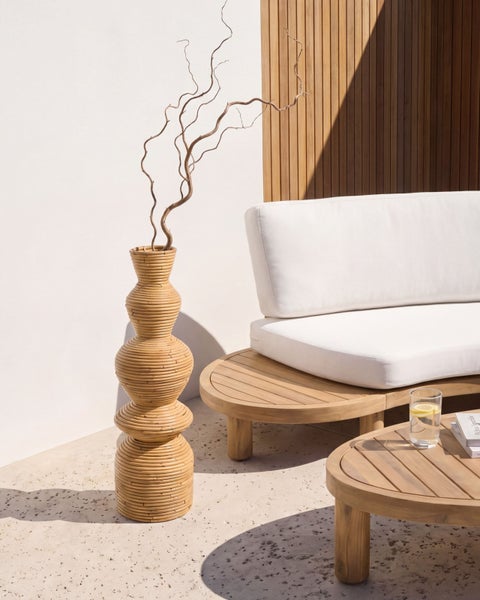 Moderner Loungebereich im Freien mit geschwungenem Holzsofa, weißen Polstern, passendem Couchtisch und einer hohen Rattanvase vor einer Holzwand.