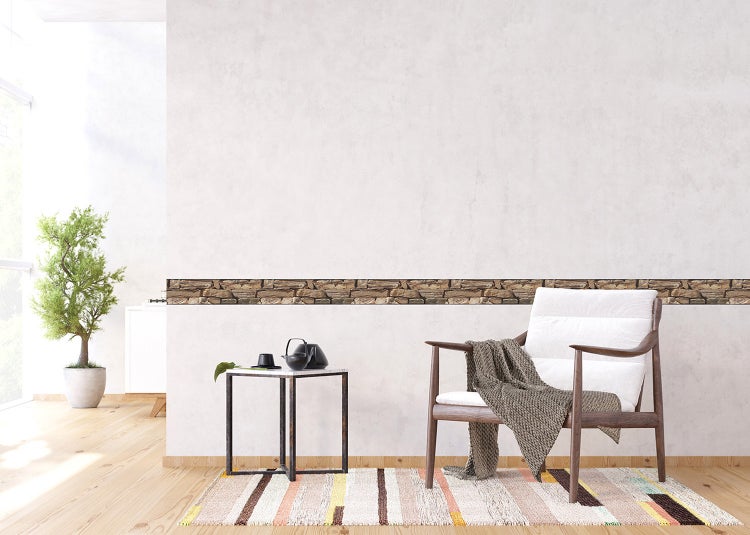 Helles Wohnzimmer mit Tapetenbordüre in Steinoptik an einer hellen Wand, einem Holzsessel und einem Beistelltisch auf einem gestreiften Teppich.