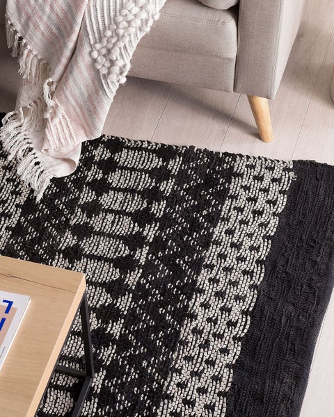 Schwarz-weißer gewebter Teppich mit geometrischem Muster in einem hellen Raum neben einem Sessel und einem Holztisch.