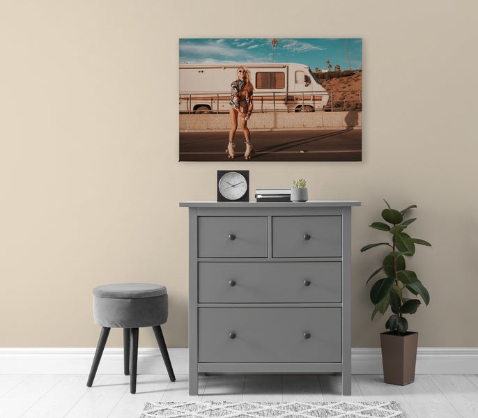Graue Kommode mit drei Schubladen unter einem Wandbild, daneben ein grauer Hocker und eine Topfpflanze.