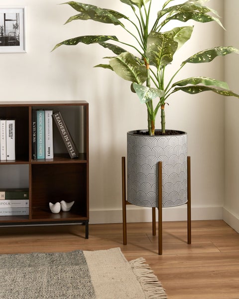 Moderner Übertopf mit geometrischem Muster auf einem Holzständer neben einem dunklen Bücherregal in einem hellen Wohnzimmer.