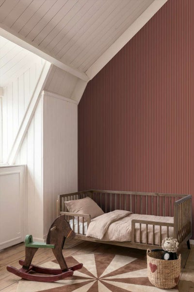 Ein Kinderzimmer mit gestreifter Tapete in Terrakotta an der Wand, einem Holzgitterbett, einem Schaukelpferd und einem runden Teppich.