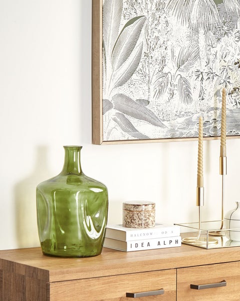 Dekorative Szene mit grüner Glasvase, Büchern, Schmuckkästchen, Kerzen und botanischem Wandbild auf einem Holzschrank