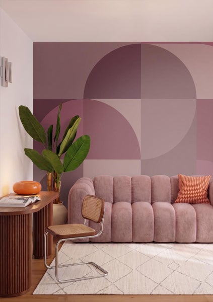 Modernes Wohnzimmer mit geometrischer Fototapete, rosa Polstersofa, Holzschreibtisch, Rattanstuhl und einer großen Zimmerpflanze.