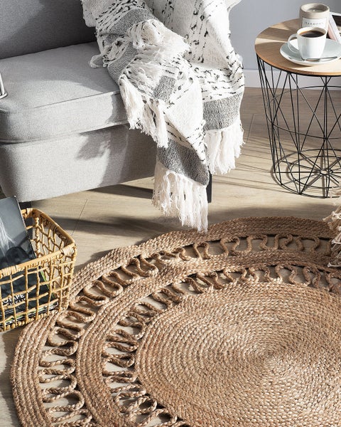 Runder gewebter Jute-Teppich mit dekorativem Schlaufenrand in einem hellen Wohnzimmer neben einem Sessel und Beistelltisch auf Holzboden.
