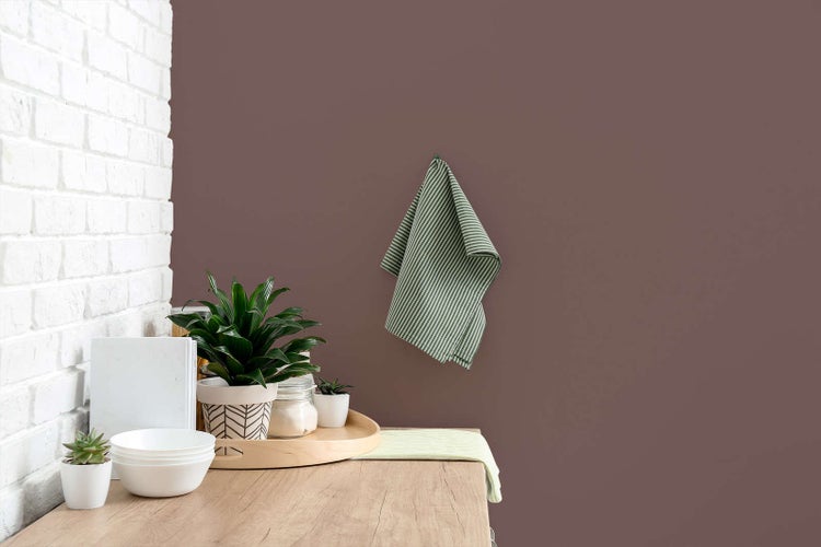 Matt violette Wand hinter einer hölzernen Arbeitsplatte mit Pflanzen, weißem Geschirr und einem gestreiften Küchentuch neben einer weißen Ziegelwand.