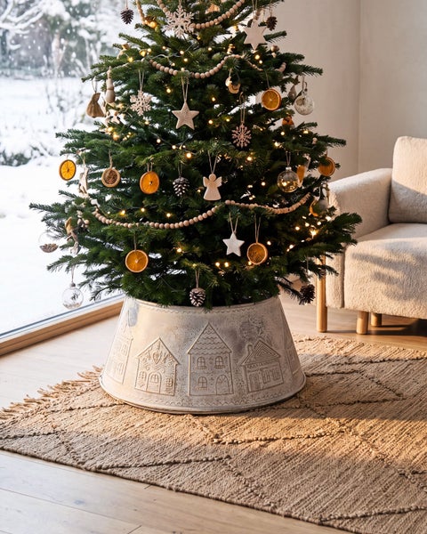 Geschmückter Weihnachtsbaum in einem runden Metall-Weihnachtsbaumständer mit eingeprägtem Häusermotiv in einem wohnlich eingerichteten Zimmer.