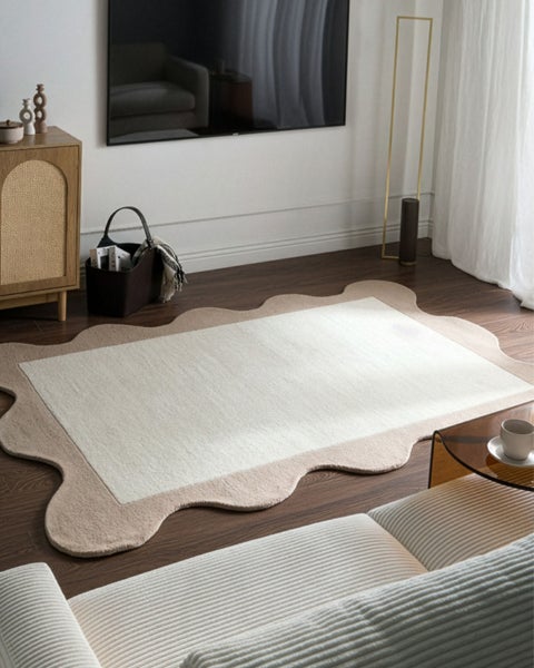 Zweifarbiger Teppich mit wellenförmigem Rand im hellen Wohnzimmer auf dunklem Holzboden neben einem Sofa.