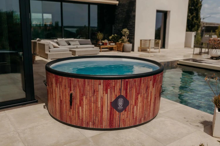 Runder aufblasbarer Whirlpool in Holzoptik auf einer modernen Terrasse neben einem Pool und Gartenmöbeln. Coco Spa Logo.
