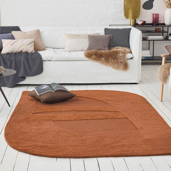 Ovaler, rostbrauner Webteppich mit geometrischem Hoch-Tief-Muster in einem hellen Wohnzimmer mit weißem Sofa und Holzfußboden.