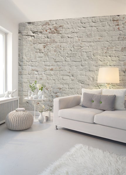 Helles Wohnzimmer mit Fototapete in rustikaler, weißer Backsteinoptik, einem weißen Sofa und einem gestrickten Hocker.