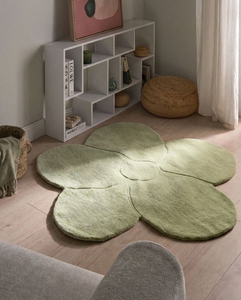 Grüner Teppich in Blumenform auf Holzboden in einem hellen Zimmer mit weißem Regal, geflochtenem Hocker und Korb.