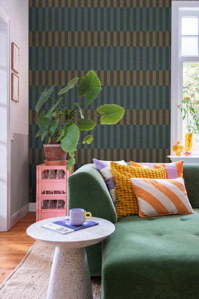 Wohnzimmer mit geometrischer Vliestapete in Dunkelgrün und Goldgelb, grünem Samtsofa, bunt gemusterten Kissen und einer großen Zimmerpflanze.