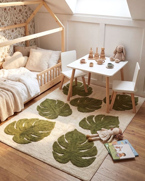 Ein Kinderzimmer mit einem Hausbett aus Holz, einem weißen Kindertisch mit Stühlen und einem Teppich mit Blattmotiv auf einem Holzboden.