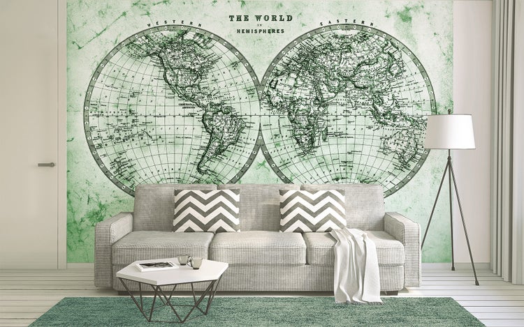 Fototapete mit historischer Weltkarte in zwei Hemisphären auf grünem Hintergrund in einem hellen Wohnzimmer hinter einem grauen Sofa.