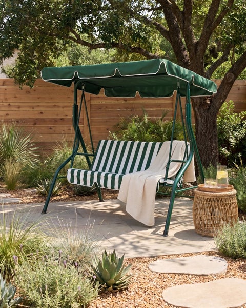 Hollywoodschaukel mit grün-weiß gestreiften Polstern, grünem Sonnendach und Metallgestell in einem Garten mit Holzschutzzaun und Pflanzen.