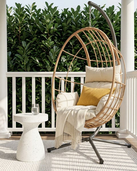 Heller Hängesessel in Ei-Form aus Geflecht mit Gestell, Kissen und Decke auf einer Terrasse mit Beistelltisch vor einer grünen Hecke.