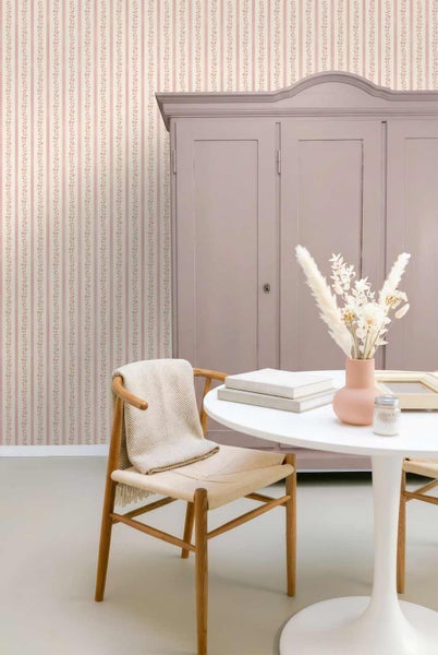 Heller Raum mit vertikal gestreifter Blumentapete, einem Holzschrank, einem weißen Rundtisch mit Trockenblumen und einem Holzstuhl.