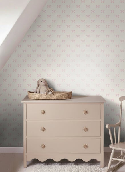 Vliestapete mit Schleifenmuster in einem hellen Kinderzimmer mit Kommode, Stofftier im Korb und Schaukelstuhl.