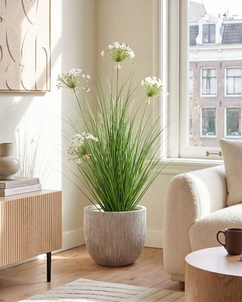 Künstliche Allium Pflanze mit hohem Gras und weißen Blüten in einem grauen strukturierten Topf in einem hellen, modernen Wohnzimmer neben einem Fenster.