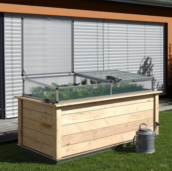Hochbeet aus Holz mit transparentem Frühbeetaufsatz und Pflanzen, eine Klappe ist geöffnet, nebenan steht eine Gießkanne auf dem Rasen.